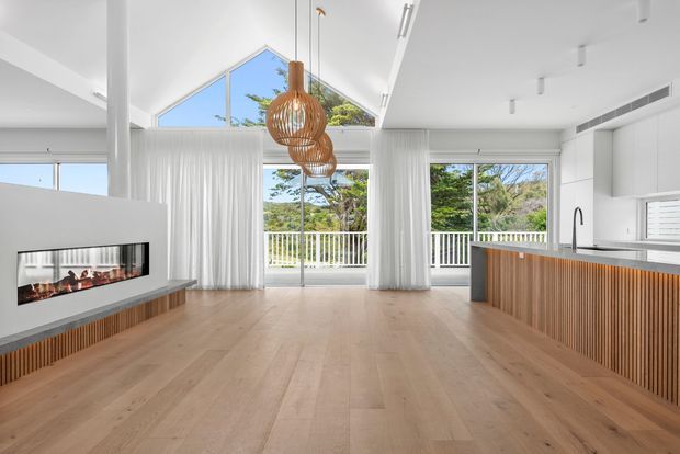 Bright open-plan living and kitchen interior with pale timber flooring, white walls and ceilings, floor-to-ceiling glazing, sheer white curtains, and a linear fireplace built into a central partition. The kitchen features a long island with vertically slatted timber cabinetry, a dark benchtop, and a black gooseneck tap. Woven timber pendant lights hang above the open space, with a balcony and greenery visible beyond the glazing.