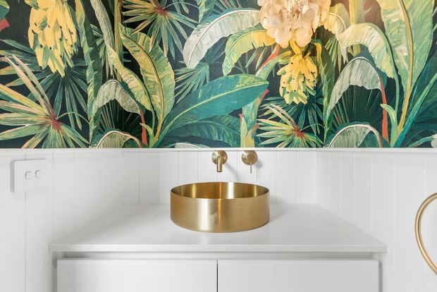 Close-up of a contemporary bathroom vanity with a white built-in cabinet and white benchtop, a round brushed-gold vessel basin and matching wall-mounted tapware. A bold tropical botanical mural forms the feature wall above the vanity, with a partial round gold-framed mirror visible at right.