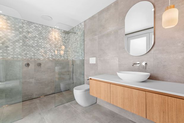 Contemporary bathroom featuring a large frameless glass walk-in shower, grey stone-look wall and floor finishes, patterned mosaic shower wall, wall-mounted rain and hand shower fittings, a white vessel basin, floating timber vanity, rounded rectangular mirror, and warm pendant lighting.