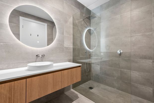 Contemporary bathroom featuring large-format grey stone-look wall and floor tiles, a floating timber vanity with a white benchtop and vessel basin, backlit round mirrors, and a frameless glass walk-in shower with recessed fixtures.