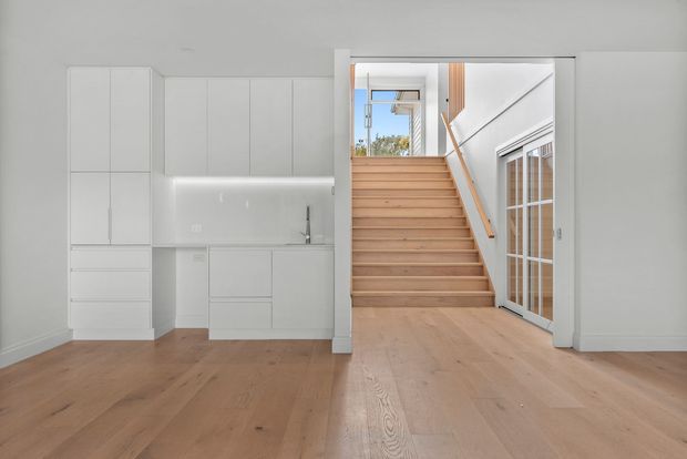 Bright, minimalist open-plan interior featuring a compact white kitchen with integrated cabinetry on the left and a timber staircase leading to an upper level at center-right. Pale timber flooring, white walls, a glass-panelled door, and abundant natural light create a clean, contemporary feel.