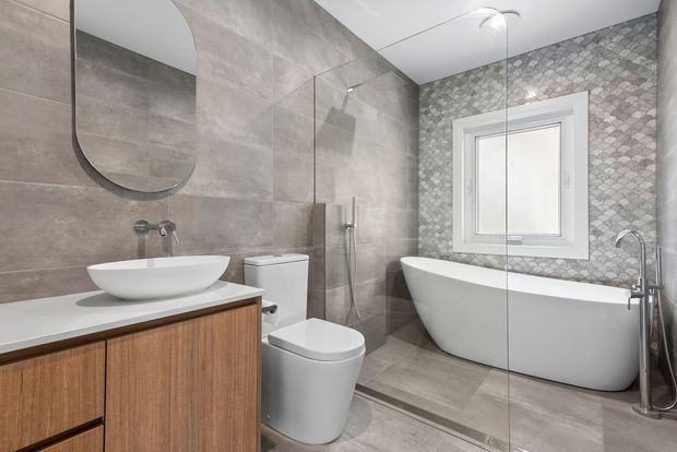 A contemporary bathroom with a freestanding white bathtub, walk-in glass shower, wall-mounted shower fixtures, toilet, and timber-veneer vanity with a vessel basin. Large-format grey wall and floor tiles are paired with a patterned grey feature wall around the window, and a rounded rectangular mirror adds a soft detail.