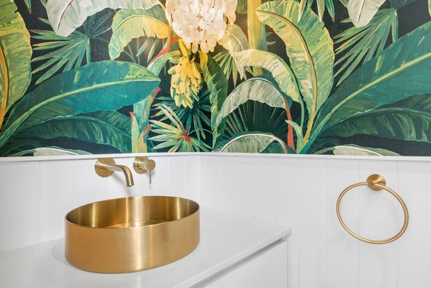 A compact bathroom vanity featuring a round brushed-gold vessel basin, matching wall-mounted tapware and towel ring, white built-in cabinetry, and white vertical wall panelling. A bold tropical botanical mural forms the feature wall above the vanity.