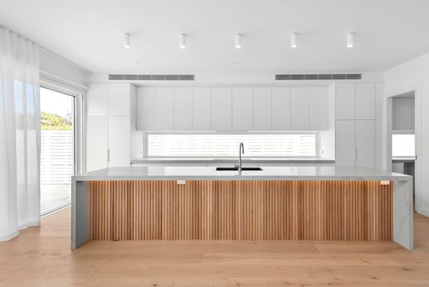 Bright contemporary kitchen with white full-height cabinetry, a large island featuring a vertically slatted timber front and grey stone benchtop, light timber flooring, integrated horizontal window, skylight-like downlights, and sheer curtains beside a glazed door.