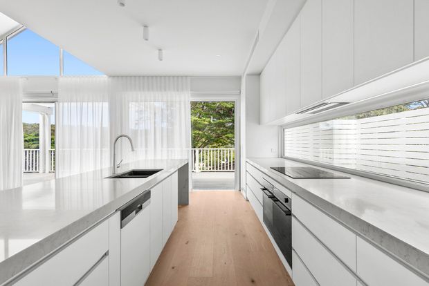 Bright contemporary white kitchen with extensive handleless cabinetry, two long stone-look benchtops, integrated stainless-steel appliances, a central island with sink, pale timber flooring, and sheer curtains opening to a balcony or outdoor area. Large windows and glazing provide abundant natural light and views of greenery.