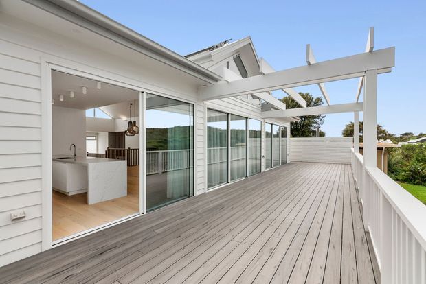 A bright, spacious timber deck and covered outdoor terrace adjoining a white weatherboard home. The deck features a white pergola with exposed rafters, white balustrades, large sliding glass doors, and views toward a green suburban landscape. Through the open doors, a contemporary kitchen with a white island and timber flooring is visible.