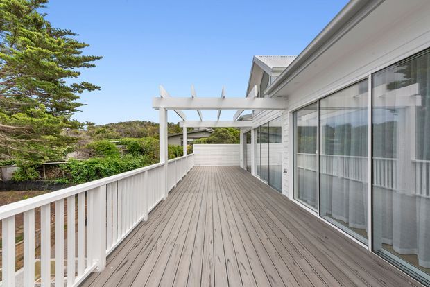 A spacious elevated outdoor deck running alongside a light-coloured contemporary house. The deck has grey timber-look decking, white vertical balustrades, a white pergola structure, and large sliding glass doors opening to the interior. Green landscaped surroundings and a clear blue sky are visible beyond the railing.