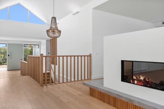 Bright contemporary open-plan interior with pale timber flooring, a timber balustrade around a stair opening, a sculptural pendant light, clerestory-style glazing, and a sleek built-in linear fireplace with a grey stone hearth and timber detailing. White walls and abundant daylight create an airy, refined atmosphere.