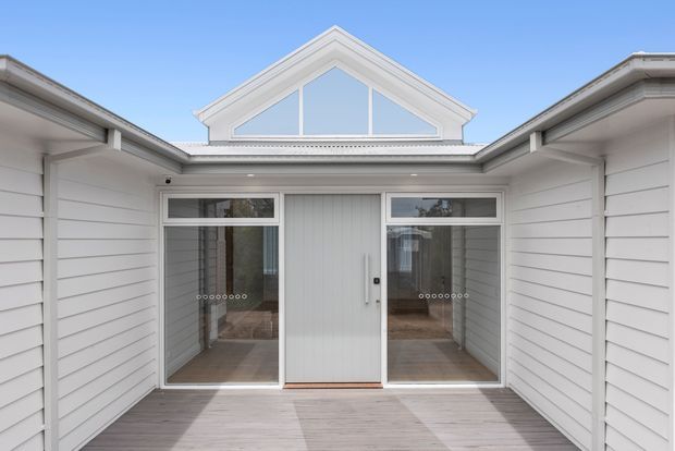 A bright white-clad courtyard or entry terrace framed by two wings of a low-rise building. The space features horizontal weatherboard walls, pale timber decking, glazed sliding doors, and a prominent triangular gable with clerestory glazing above the central entrance. The composition is clean and symmetrical under a clear blue sky.
