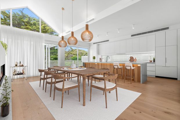 Bright open-plan dining and kitchen interior with a large timber dining table and matching chairs set on a light textured rug. The white kitchen features full-height cabinetry, a long island with a timber slatted base, and integrated appliances. Floor-to-ceiling glazing and sheer curtains open toward greenery, while sculptural woven pendant lights provide warm accents. Light timber flooring and abundant daylight create an airy, contemporary atmosphere.