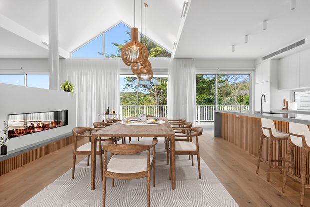 Bright open-plan dining and kitchen interior with a timber dining table and chairs, a white kitchen with timber island cabinetry, a built-in linear fireplace, floor-to-ceiling glazing, and a high triangular clerestory window. Natural light, sheer curtains, timber finishes, and woven pendant lights create a warm contemporary coastal atmosphere.