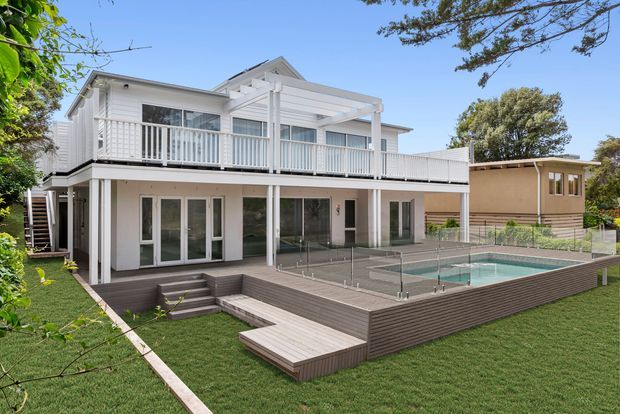 Exterior view of a bright two-storey residential home with white weatherboard-style cladding, extensive upper-level balcony and pergola, glass doors, and a raised timber-look deck surrounding a fenced swimming pool. The landscaped backyard includes lawn and mature trees, with a neighbouring low-rise structure visible beyond the property.