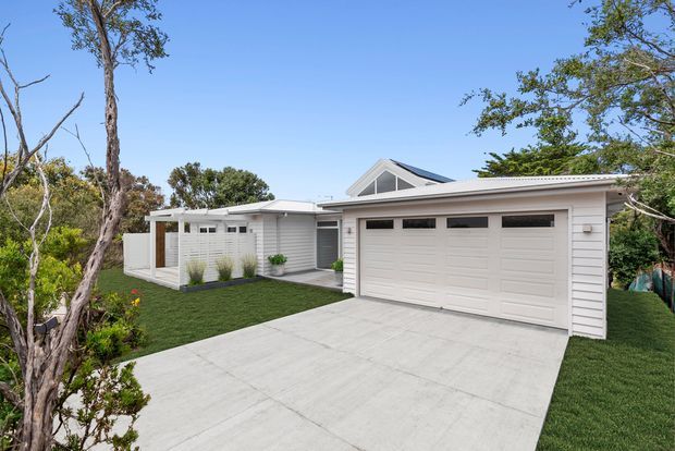 Contemporary single-storey suburban home exterior with a broad white sectional garage door, concrete driveway, pale horizontal cladding, a low flat-roofed entry wing, and a pitched gable roof visible behind. The house is set among established trees and landscaped lawn.