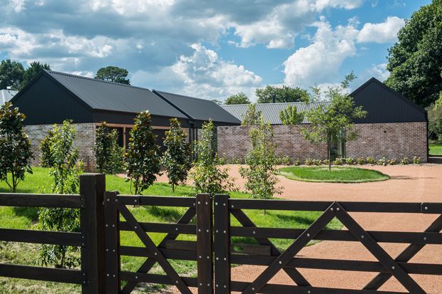 Exterior view of a low-rise rural or semi-rural property composed of several connected gabled buildings. The structures combine dark corrugated metal roofing and dark gable cladding with exposed warm brick walls and timber-framed openings. A timber farm-style gate and fence occupy the foreground, with a landscaped courtyard, young trees, gravel driveway, and mature vegetation around the buildings. Bright daylight and dramatic clouds create a crisp, inviting setting.