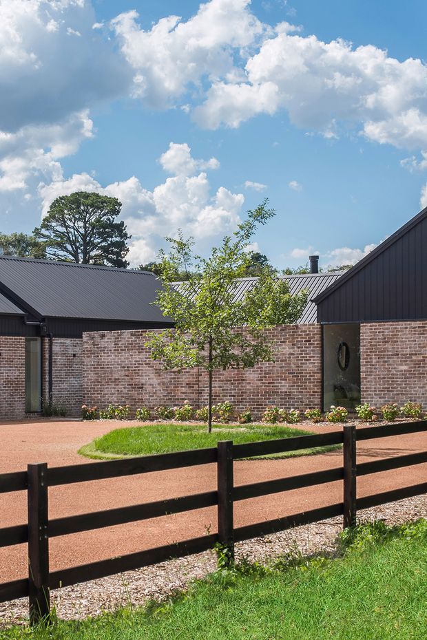 Exterior view of a low-rise property with dark metal gabled roofs, dark vertical cladding, and extensive exposed red brick walls. A landscaped courtyard or forecourt includes a central young tree, planted garden beds, reddish gravel or paved paths, and a black timber post-and-rail fence in the foreground. Bright blue sky and mature trees form the background.