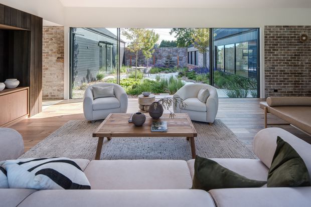 A bright contemporary living room with large floor-to-ceiling glazing opening onto a landscaped courtyard. The room features exposed brick walls, timber built-in cabinetry, pale upholstered sofas and armchairs, a timber coffee table, and a neutral textured rug. The indoor-outdoor connection and courtyard garden dominate the view.