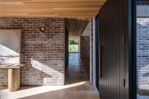 A sunlit residential entry passage with exposed brick walls, large beige stone or porcelain floor tiles, warm timber-lined ceiling, and a tall dark vertical-panel door beside black-framed glazing. The corridor leads toward a small outdoor garden view, creating a strong indoor–outdoor connection. Built-in and architectural finishes dominate, with simple contemporary detailing and rustic brick texture.