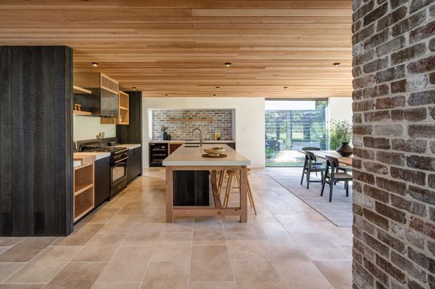 Open-plan kitchen and dining area with a central timber island, dark wood cabinetry, exposed brick feature walls, a warm timber-lined ceiling, and large glazed doors opening to the outdoors. The space has a contemporary, earthy character with abundant natural light.
