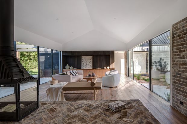A bright contemporary living room with a dramatic vaulted ceiling, extensive floor-to-ceiling glazing, and timber flooring. The room features a central seating arrangement with pale upholstered sofas and armchairs, a low timber coffee table, a dark timber feature wall with built-in joinery, and an exposed brick wall. A large black sculptural fireplace or stove occupies the left foreground, while a patterned area rug defines the seating zone. The glazing opens the room toward a landscaped courtyard or garden.
