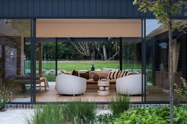 A contemporary living room viewed from the landscaped exterior through expansive floor-to-ceiling glazing. The room features a low sculptural seating arrangement, rounded cream lounge chairs, a tan sofa with striped cushions, a stacked stone or concrete coffee table, timber flooring, dark-framed glazing, and exposed brick at the side. The space opens directly onto a lush garden and lawn, creating a strong indoor–outdoor connection.