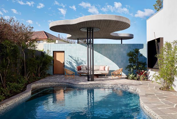 A sculptural outdoor pool courtyard with a curved concrete canopy supported by slender dark columns, positioned over an outdoor lounge area. The pool has a curved tiled edge, while irregular stone paving surrounds the space. A rendered wall, timber door, planting, and a modern white building frame the private setting under a bright blue sky.