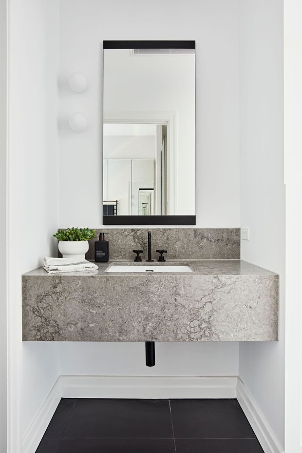 A compact, modern bathroom vanity in a narrow white alcove. The wall-mounted vanity and integrated basin are clad in grey veined stone, paired with a matte black wall-mounted tap set and a large rectangular black-framed mirror. White walls and trim contrast with dark floor tiles, while a small plant and soap dispenser sit on the benchtop.