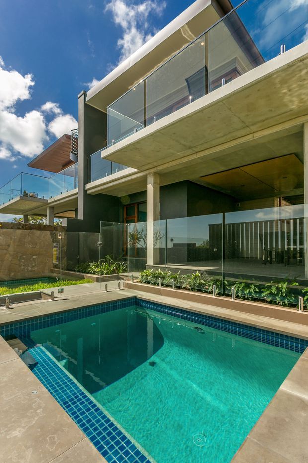 Exterior view of a multi-level contemporary home with broad concrete cantilevered balconies, dark rendered walls, extensive clear glass balustrades, and a ground-level covered terrace. A rectangular tiled swimming pool dominates the foreground, with landscaped planting and paved surrounds. Bright blue sky and strong sunlight emphasize the open, resort-like setting.