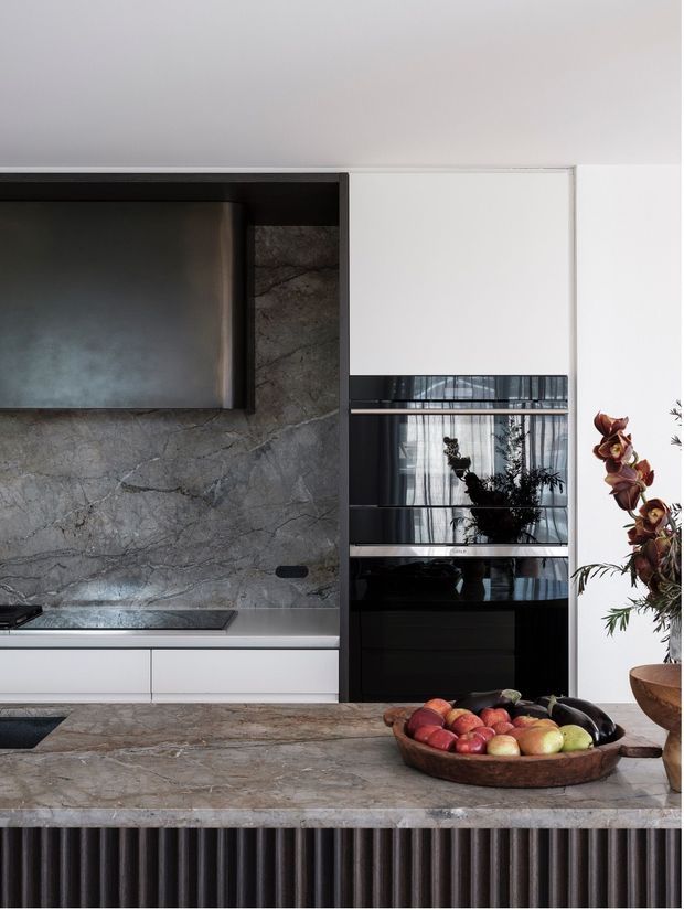 A sophisticated contemporary kitchen featuring dark grey-veined stone across the backsplash and island, integrated white cabinetry, a sculptural dark metal rangehood, and a black-framed glazed display or pantry unit. The island has a richly textured stone surface with a fluted dark base, complemented by restrained styling and warm natural produce.