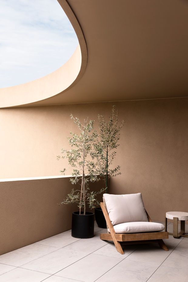 A serene, minimalist indoor-outdoor lounge nook with warm taupe plaster walls, a dramatic curved ceiling opening bringing in daylight, large pale floor tiles, and a timber-framed lounge chair with a light cushion. Potted olive trees and a small side table complete the calm, earthy setting.
