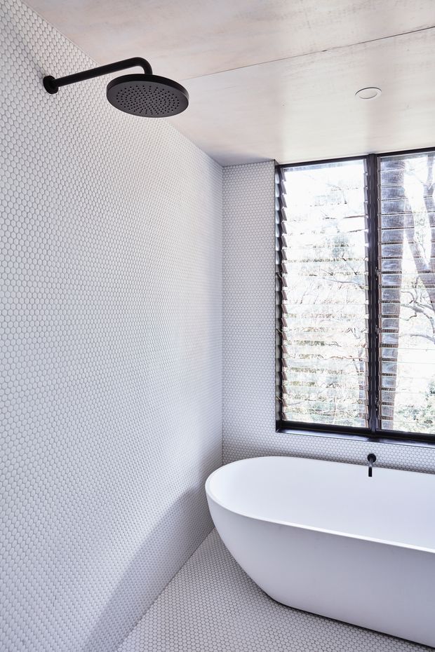 Bright contemporary bathroom featuring a freestanding white bathtub, wall-mounted matte black round shower head and tap, continuous small-format white hexagonal tile across the walls and floor, and a large black-framed louvred window overlooking trees. The space is clean, minimal, and naturally lit.