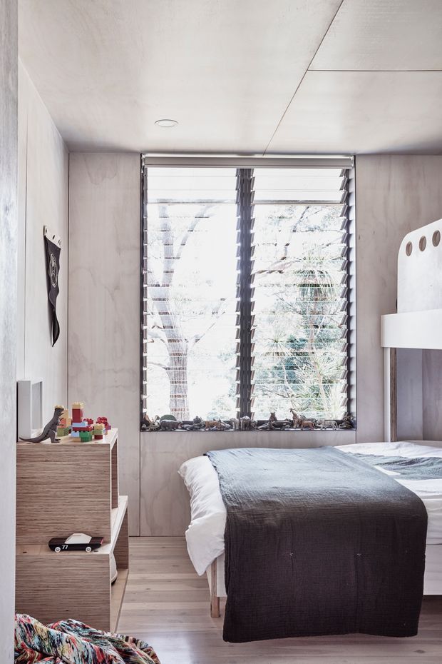 A compact, contemporary bedroom with pale timber wall panelling and matching timber flooring. A low bed is dressed in white and charcoal-grey bedding, positioned in front of a large louvred window overlooking trees. Built-in stepped storage sits along the left wall, displaying small toys and objects, while a white wall-mounted shelf is visible on the right. The room has a restrained neutral palette, abundant filtered daylight, and a clean, functional built-in design.