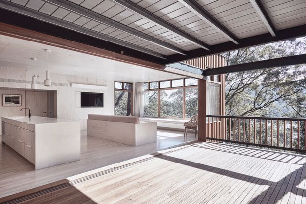An airy, open-plan contemporary living and kitchen space opening onto a large timber deck. The room features a white kitchen island with integrated cabinetry, pale timber flooring, a streamlined white kitchen and cabinetry, broad timber-framed glazing, and a partially covered deck with dark structural beams and railing. Surrounding trees are visible beyond the windows and deck.