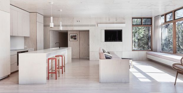 Bright open-plan contemporary interior combining a kitchen, dining, and living area. Pale timber-toned cabinetry and wall finishes, a large white island with red stools, integrated appliances, built-in living-room storage and seating, and expansive timber-framed windows define the space. The room has a clean, light-filled minimalist appearance with views to trees outside.