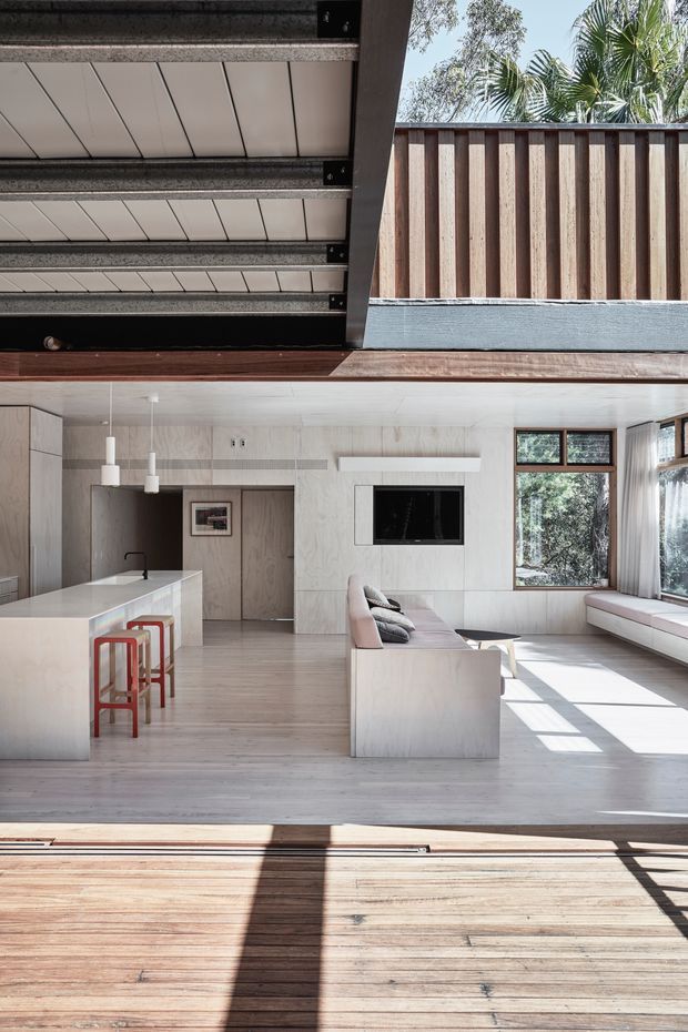 A bright, minimalist open-plan living room and kitchen viewed from a timber deck through a large opening. The interior features pale timber wall and floor finishes, a white island bench with red bar stools, a streamlined sofa, built-in window seating, pendant lights, and large windows. Above, a partially open or glazed roof structure and timber privacy screen are visible, with strong sunlight casting shadows across the deck and floor.