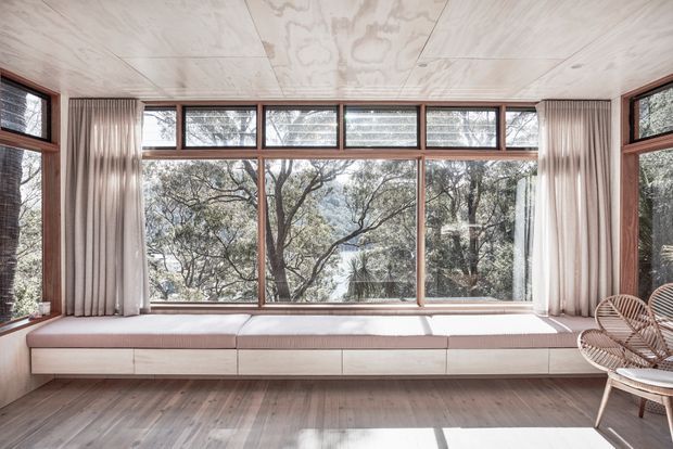 A bright, serene sunroom with expansive timber-framed glazing overlooking trees. A built-in upholstered window seat spans the room beneath the windows, complemented by sheer curtains and woven rattan lounge chairs. The room features a pale plywood-lined ceiling, warm timber flooring, and a soft neutral palette.