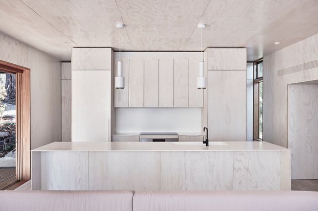 A bright, minimalist kitchen viewed from behind a sofa or seating area. The space features a large pale island with an integrated light benchtop, full-height pale timber cabinetry, matching timber-lined walls and ceiling, slim pendant lights, and a black tap. Tall glazing and a timber-framed opening bring in natural light and views of the outdoors.
