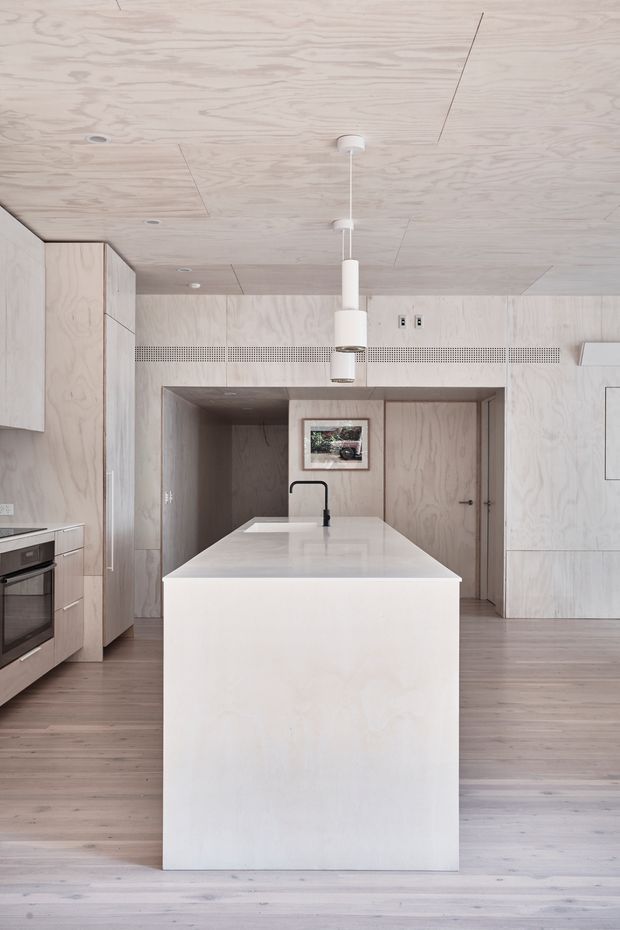 Minimal, light-toned kitchen centred on a large island with an integrated sink and matte black tap. Pale plywood cabinetry and wall panels extend across the room, complemented by wide timber flooring, a built-in oven, concealed storage, and simple white cylindrical pendant lights. A perforated ventilation grille runs horizontally across the rear wall, with a doorway and framed artwork visible beyond the island.