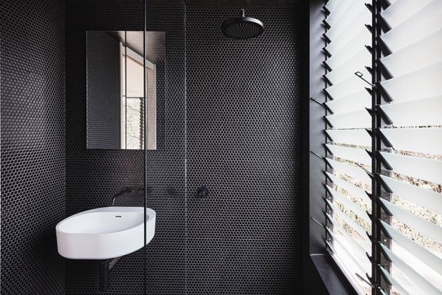 A compact, dramatically styled bathroom with black hexagonal mosaic wall and floor tiles, a white wall-mounted basin, black fixtures, a frameless glass shower enclosure, and a large louvred window admitting bright daylight. The space has a strong monochrome, contemporary-industrial character.