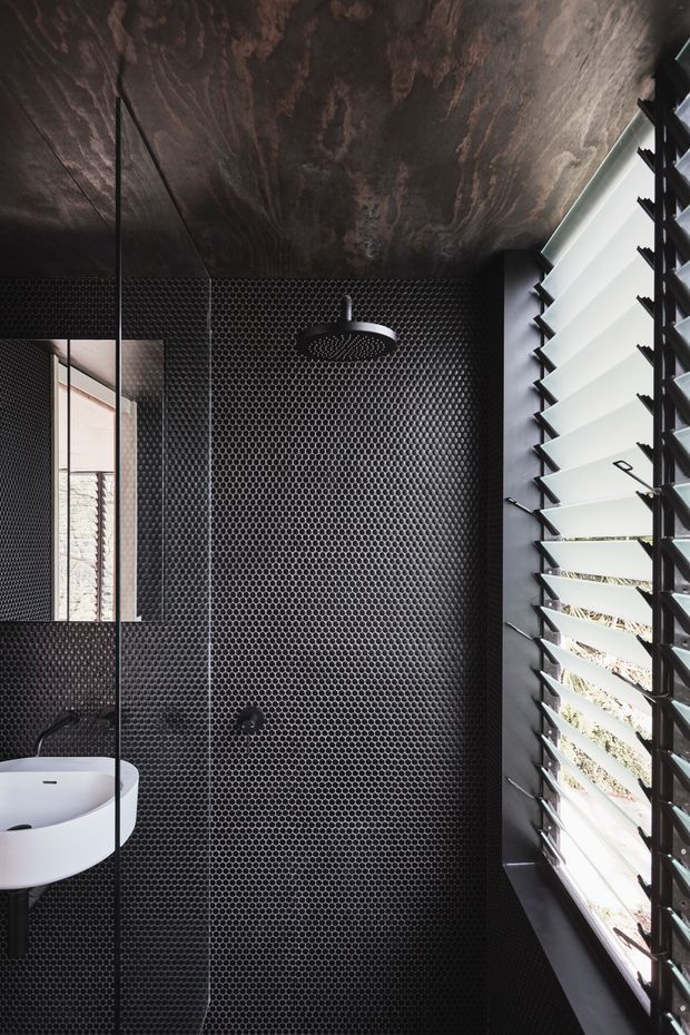 A compact, dramatic contemporary bathroom with black small-format hexagonal tiles covering the shower walls and floor, a dark richly textured timber ceiling, and a white wall-mounted basin visible beside a frameless glass shower screen. A large black-framed louvred window brings in strong daylight and views of exterior planting. The palette is predominantly black, charcoal, white, and dark timber, creating a moody high-contrast interior.