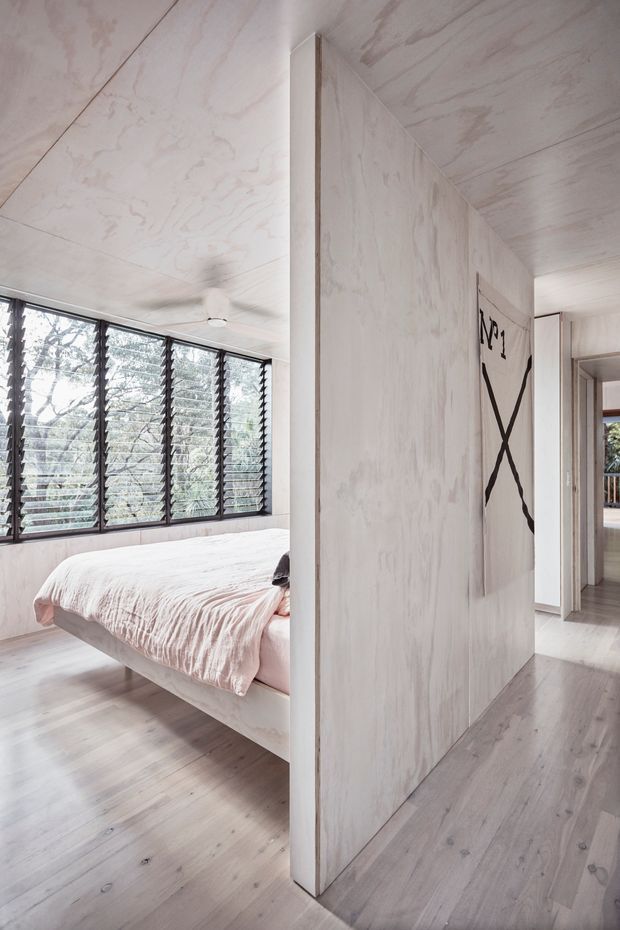 Bright, minimalist bedroom viewed from a central partitioned hallway. The room features a low platform bed with pale pink bedding, pale timber flooring, whitewashed timber wall and ceiling panels, and a broad bank of dark-framed louvre windows overlooking trees. A simple monochrome artwork hangs on the partition wall, with a glimpse of another interior space beyond the doorway.