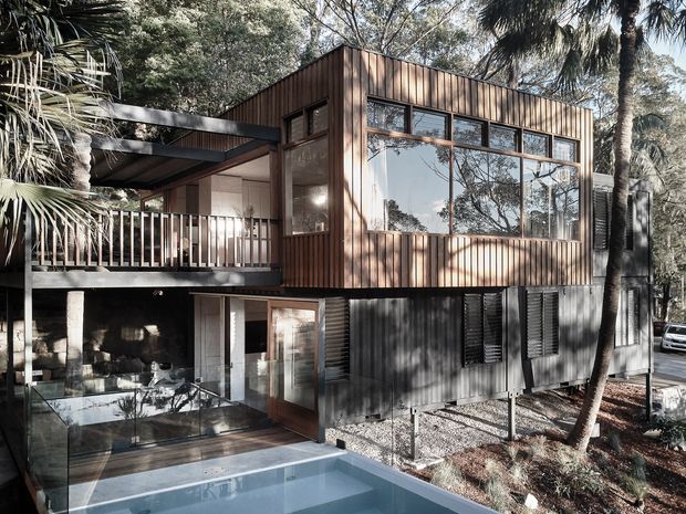 A contemporary multi-level house nestled among mature trees, viewed from the exterior. The upper volume is clad in warm vertical timber boards with extensive horizontal glazing, while the lower level uses dark corrugated metal cladding and operable louvred shutters. A covered balcony and outdoor entertaining area overlook a compact swimming pool, with the house elevated above a landscaped, wooded site.