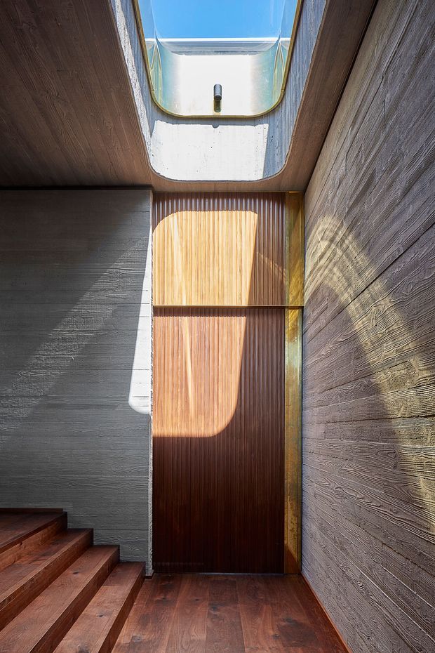 A narrow, sculptural interior stair hall with dark timber flooring and stepped timber stairs at left. The space is enclosed by heavily textured grey concrete or plaster walls, with a tall vertical timber slat feature and door at the end. A large overhead skylight with a curved concrete reveal brings in strong sunlight, creating dramatic geometric shadows. The design has an austere, material-focused contemporary character.