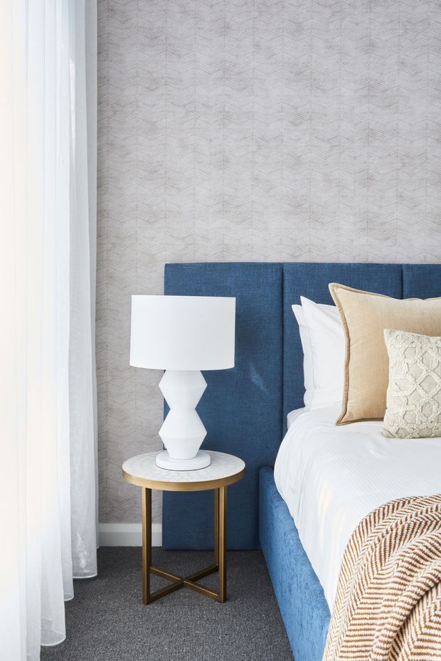 Bright, refined bedroom vignette featuring a deep blue upholstered bed and tall headboard, crisp white bedding, layered beige and cream decorative pillows, and a small round bedside table with a white sculptural lamp and brass-toned legs. A subtle geometric patterned wallpaper forms the backdrop, with sheer curtains at the window and dark grey carpet underfoot.