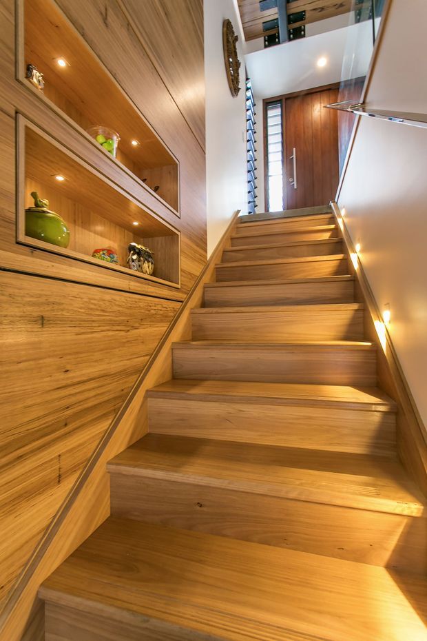A warm, timber-lined interior stairway ascending toward a wooden entry door. The staircase has broad solid-wood treads and integrated timber side walls, with built-in open display shelving and cabinetry along the left wall, recessed lighting, and a white plaster wall on the right. A narrow vertical window brings additional daylight into the stair hall.