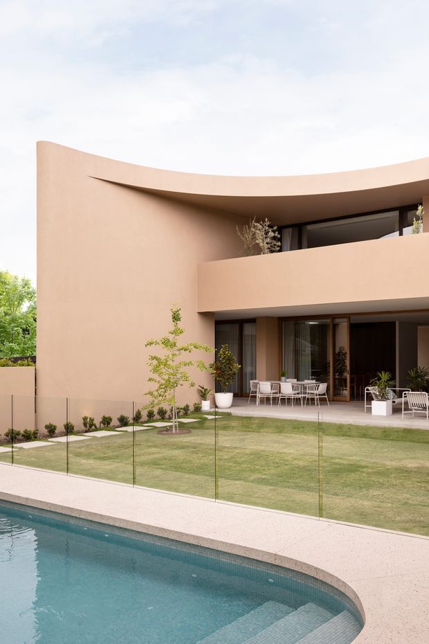 Modern two-storey residential home with a curved rendered facade and deep roof overhang, opening onto a landscaped lawn and outdoor entertaining terrace. Floor-to-ceiling glazing, timber-framed doors, minimalist outdoor furniture, frameless glass pool fencing, and a curved swimming pool define the scene.