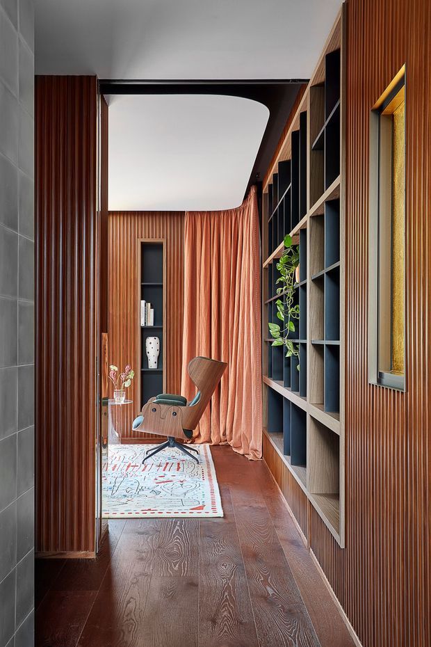 A narrow, sophisticated interior corridor or study nook featuring dark timber flooring, extensive vertical timber wall panelling, built-in open shelving, and a sculptural mid-century-style lounge chair. A curved ceiling opening frames a softly draped coral curtain at the rear, while a patterned rug, small side table, books, vase, and trailing plant add character. The space has a warm, refined, richly layered contemporary design.
