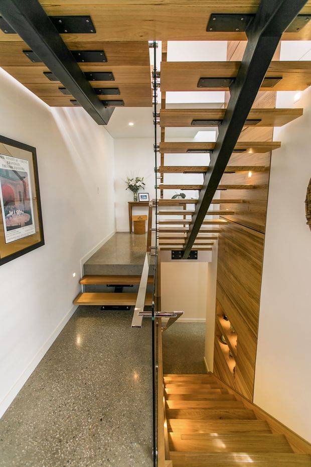 Interior view of a contemporary stairway featuring open-riser timber stairs, black steel structural supports, a clear glass balustrade, polished concrete or terrazzo flooring, and a timber-lined stairwell wall with integrated lighting. The stair connects multiple levels in a narrow, tall circulation space.