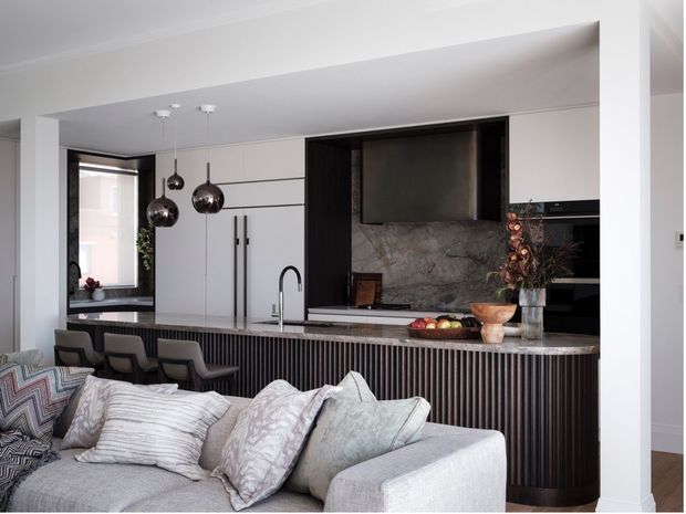 Open-plan contemporary kitchen viewed from the living area, featuring a large stone island with integrated sink, dark fluted cabinetry, white full-height cabinetry, marble-look splashback, pendant lights, and a grey upholstered sofa in the foreground.