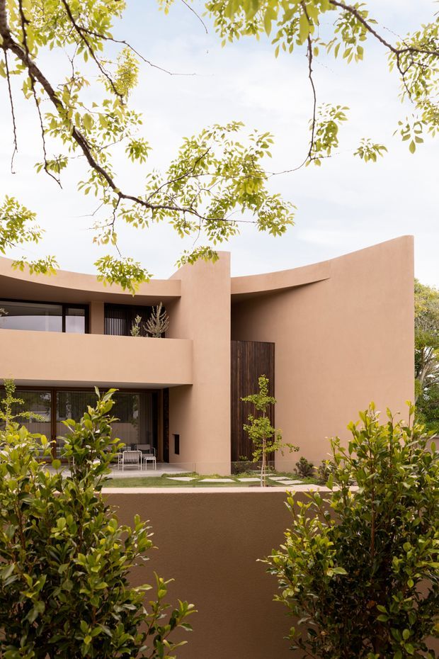 Exterior view of a contemporary two-storey residence with a sculptural curved parapet and rounded facade forms, rendered in warm terracotta tones. Dark vertical timber screening, expansive glazed openings, recessed balconies, and minimalist landscaping frame the building.