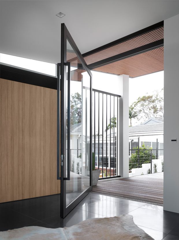 A contemporary residential entryway opens through a tall black-framed glazed door onto a timber deck or covered outdoor area. The interior features a large vertical timber wall panel, glossy dark flooring with a pale hide rug, white walls, and a slatted timber ceiling extending outdoors. Black metal balustrades and nearby white-roofed homes are visible beyond the opening.